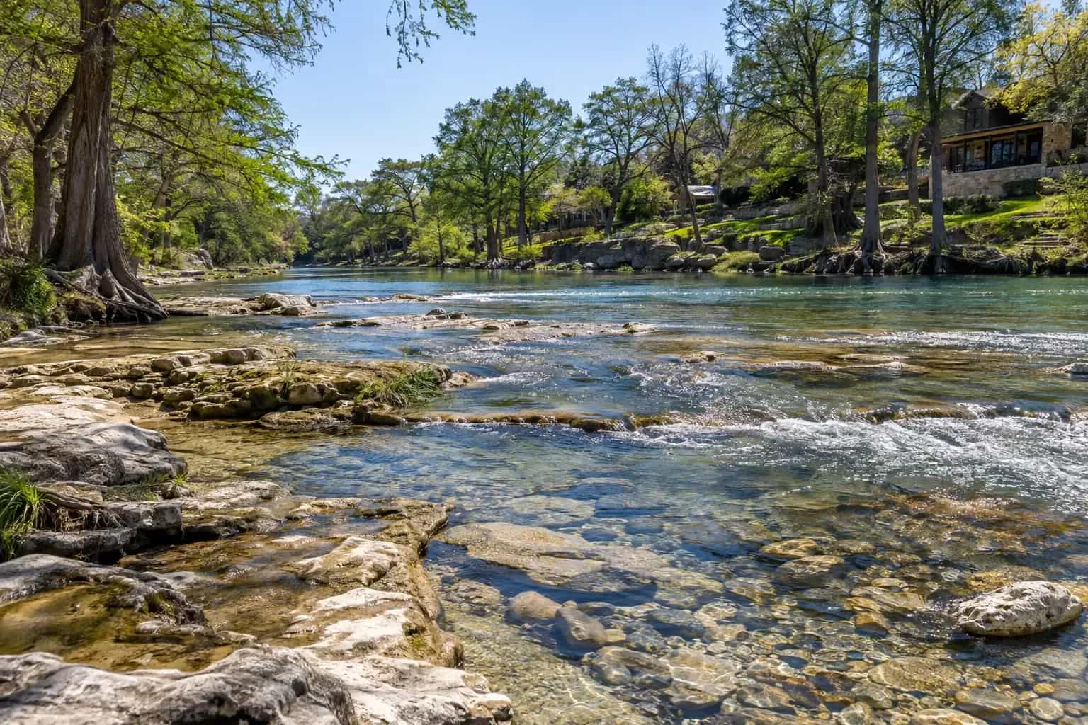 The Guadalupe River running over a limestone bed past cypress trees and riverside homes in New Braunfels, TX