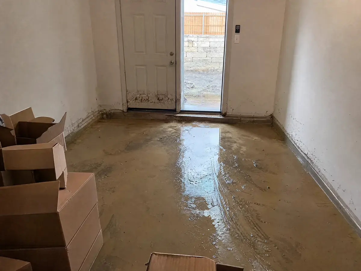 Converted garage room in a newer subdivision home with muddy storm runoff across the floor, a silt trail from the exterior door and boxes wicking water.