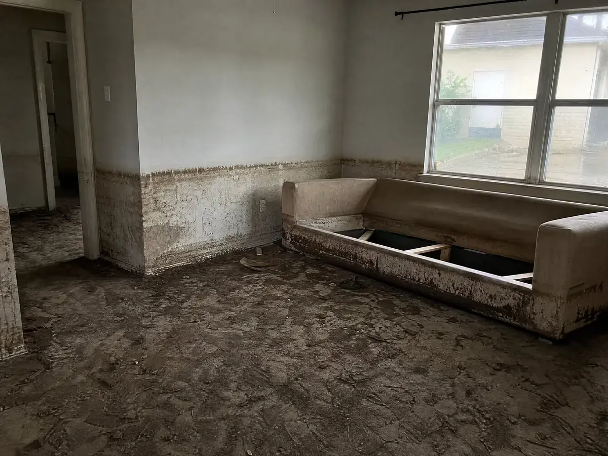 Living room after river flooding, with a muddy brown flood line two feet up the wall, silt across the floor and a stained sofa base.