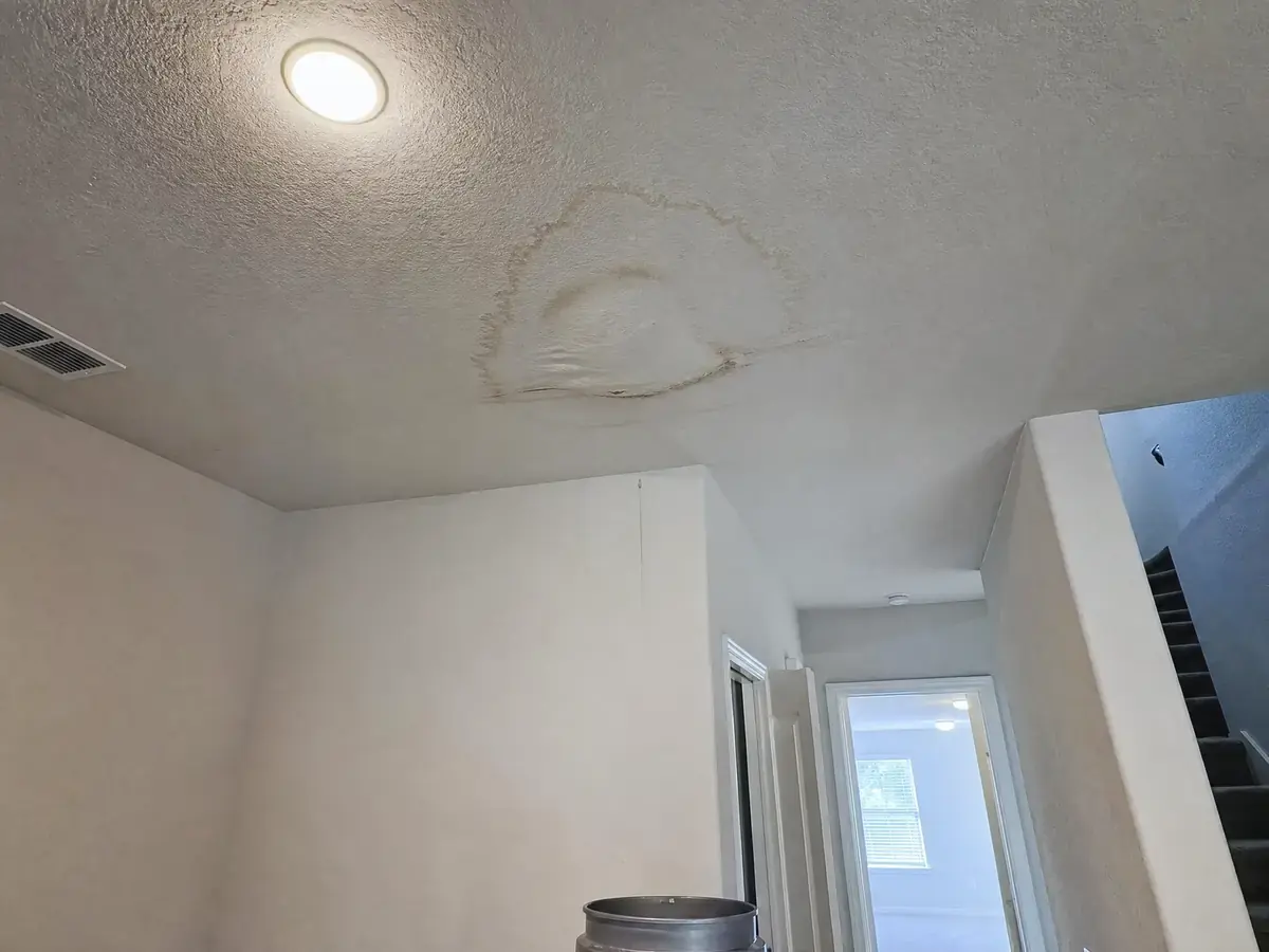 Downstairs ceiling below an upstairs bathroom with a brown water stain and dark ring, bulging drywall and a bucket on the floor underneath.