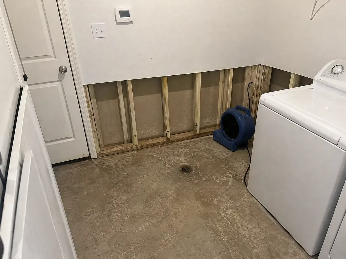 The same utility room stripped to clean slab, drywall cut out to 24 inches with treated studs exposed and an air mover running in the corner.