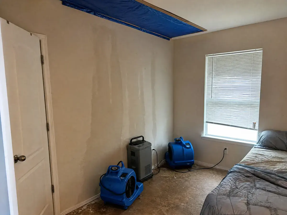 The same bedroom with the ceiling opening sealed from inside by a taut tarp, wet carpet removed to subfloor and drying equipment running.