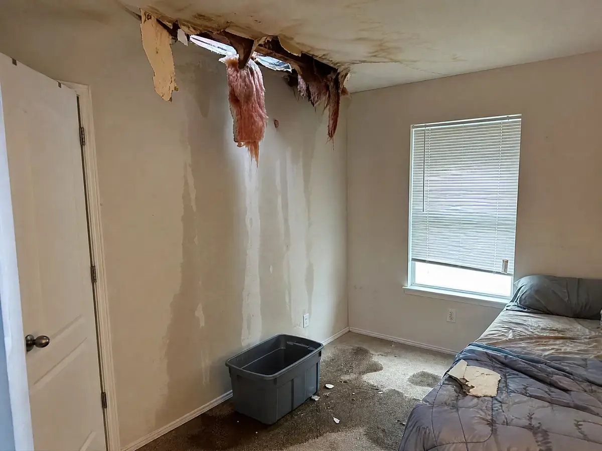Bedroom ceiling torn open by storm damage with wet insulation hanging through, daylight visible above and a soaked patch on the carpet below.