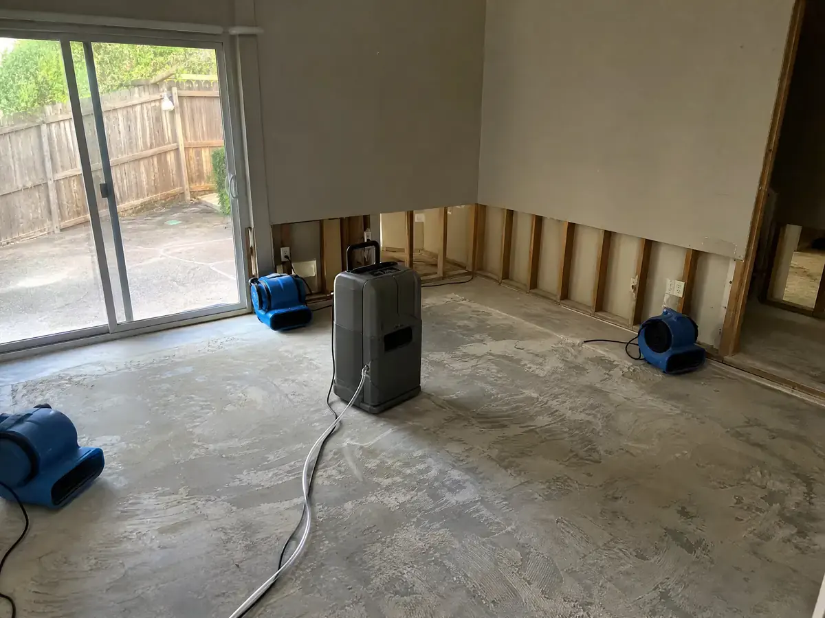 Living room stripped to bare slab with a flood cut along the wall, air movers and an LGR dehumidifier running on day one.