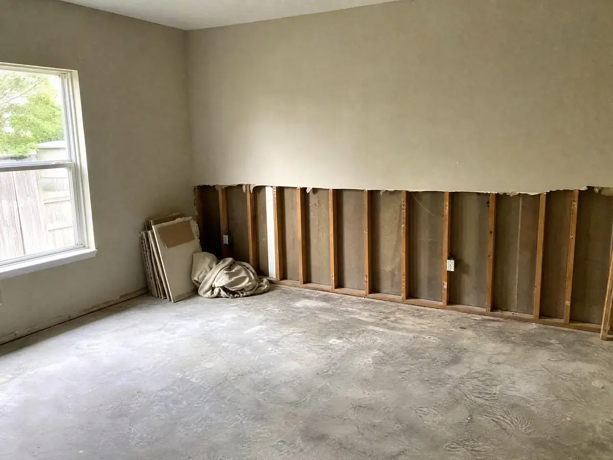 Empty bedroom mid-restoration with the lower half of one wall cut away, exposed studs and empty insulation bays, and bare concrete slab underfoot.