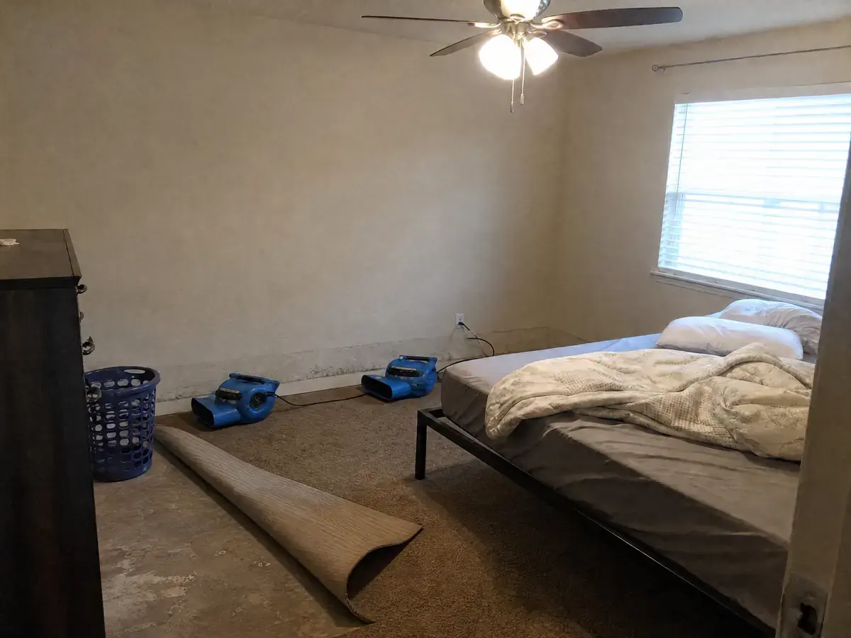 The same bedroom with the standing water gone, carpet folded back off the slab and air movers running along the wet wall line.