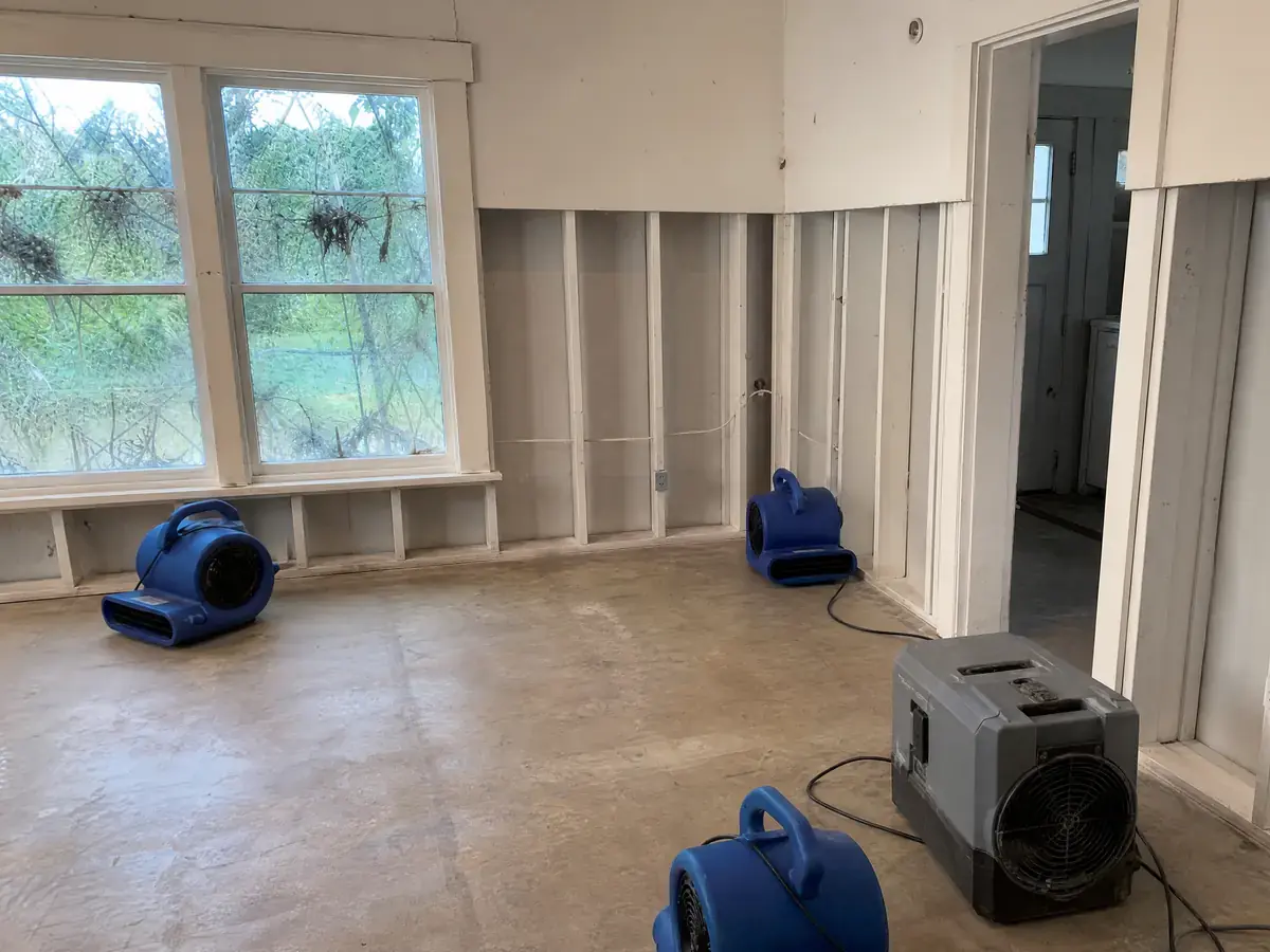 The same room stripped to clean subfloor with drywall cut well above the old flood line, treated framing exposed and three air movers running.