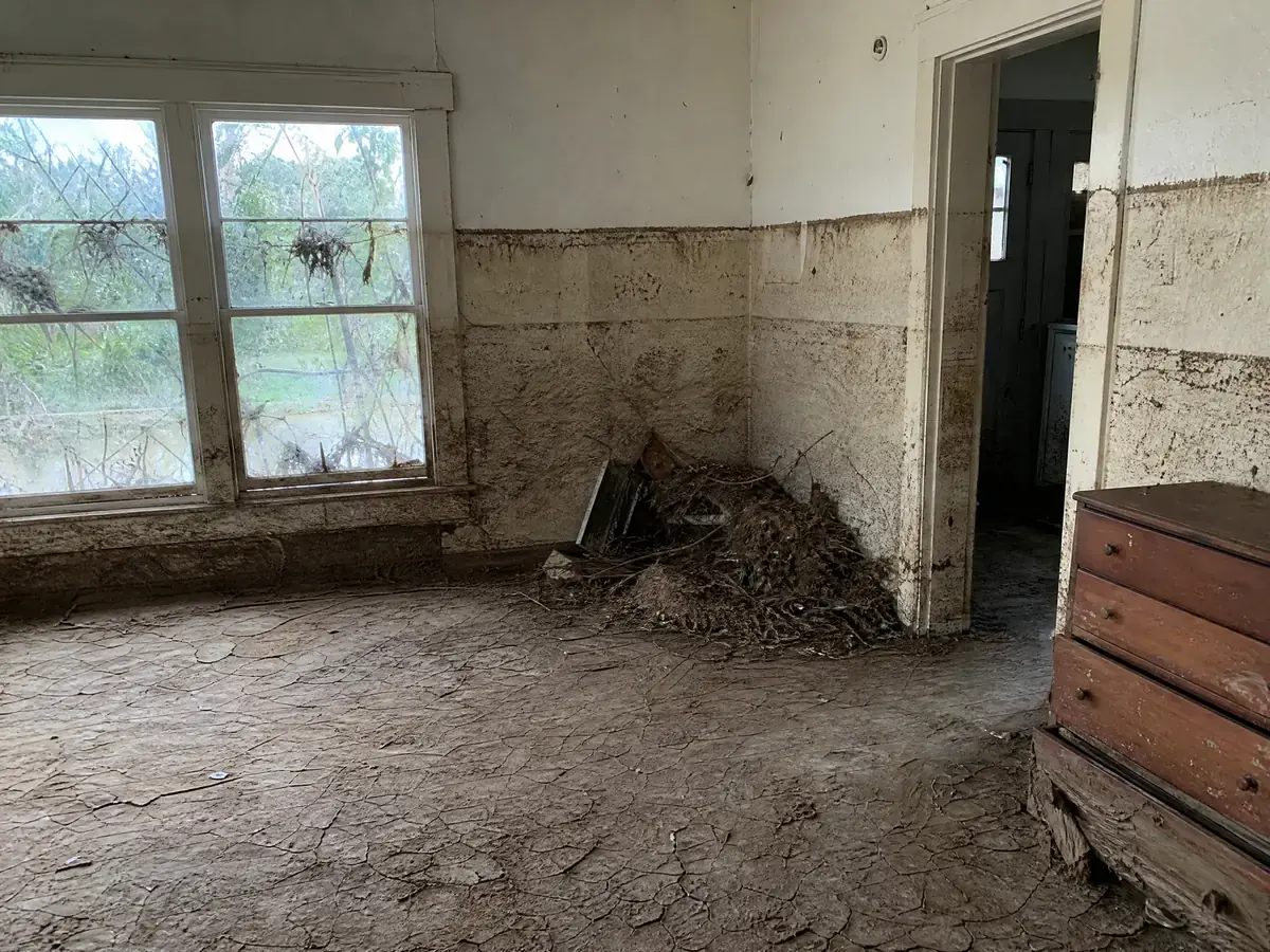 Hill Country living room after river flooding, with a heavy mud line more than three feet up the wall, dried cracked silt on the floor and river debris in the corner.